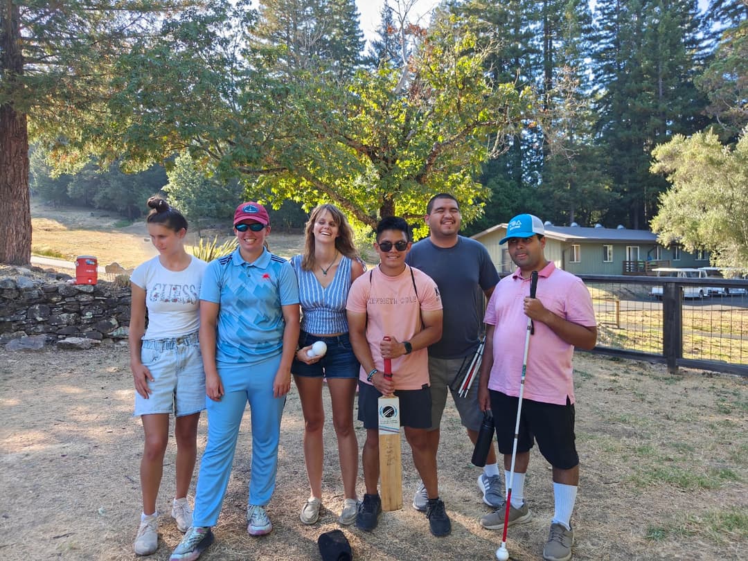 A group of 3 visually impaired female and 3 visually impaired men posing for the camera while a man holding a bat and woman holding cricket balls.