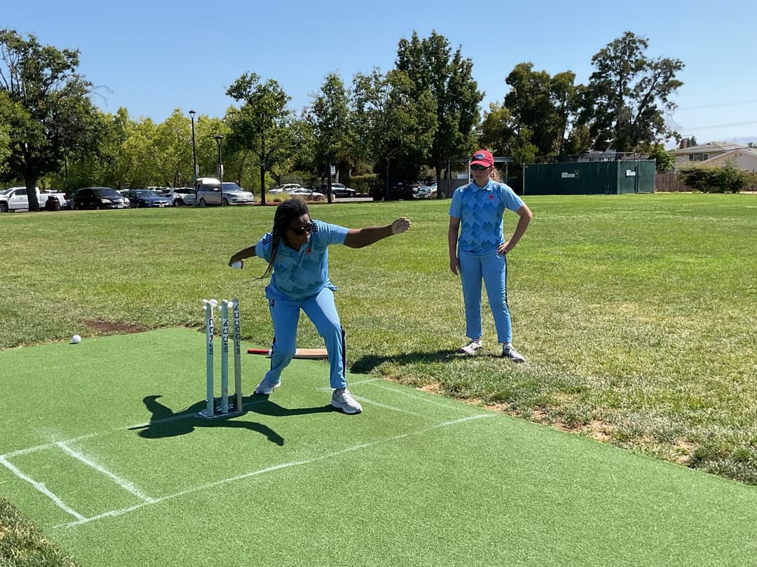 A bowler in the action to bowl, the ball has left her hand. The non striker, couple of fielders and the umpire are also visible in the shot. 