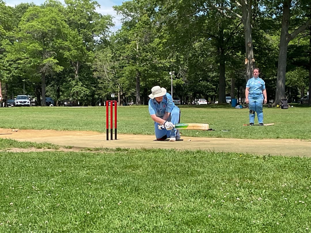 A bowler in the action to bowl, the ball has left her hand. The non striker, couple of fielders and the umpire are also visible in the shot. 