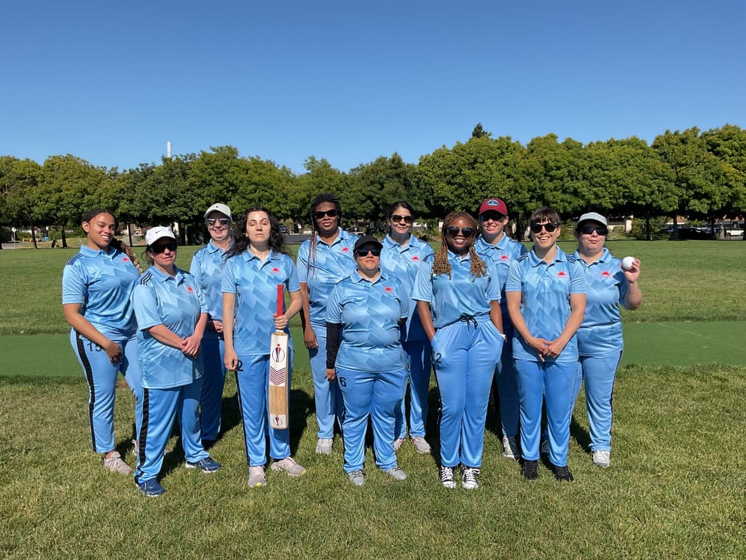 A bowler in the action to bowl, the ball has left her hand. The non striker, couple of fielders and the umpire are also visible in the shot. 