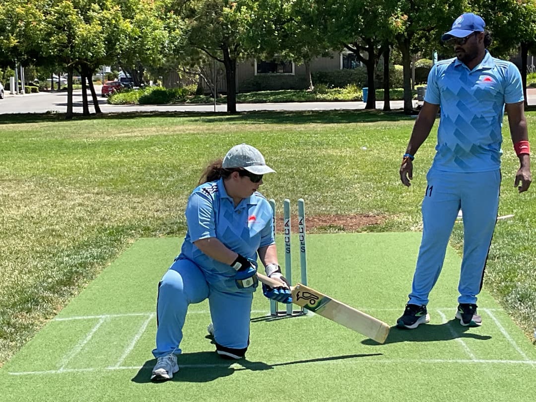 A bowler in the action to bowl, the ball has left her hand. The non striker, couple of fielders and the umpire are also visible in the shot. 