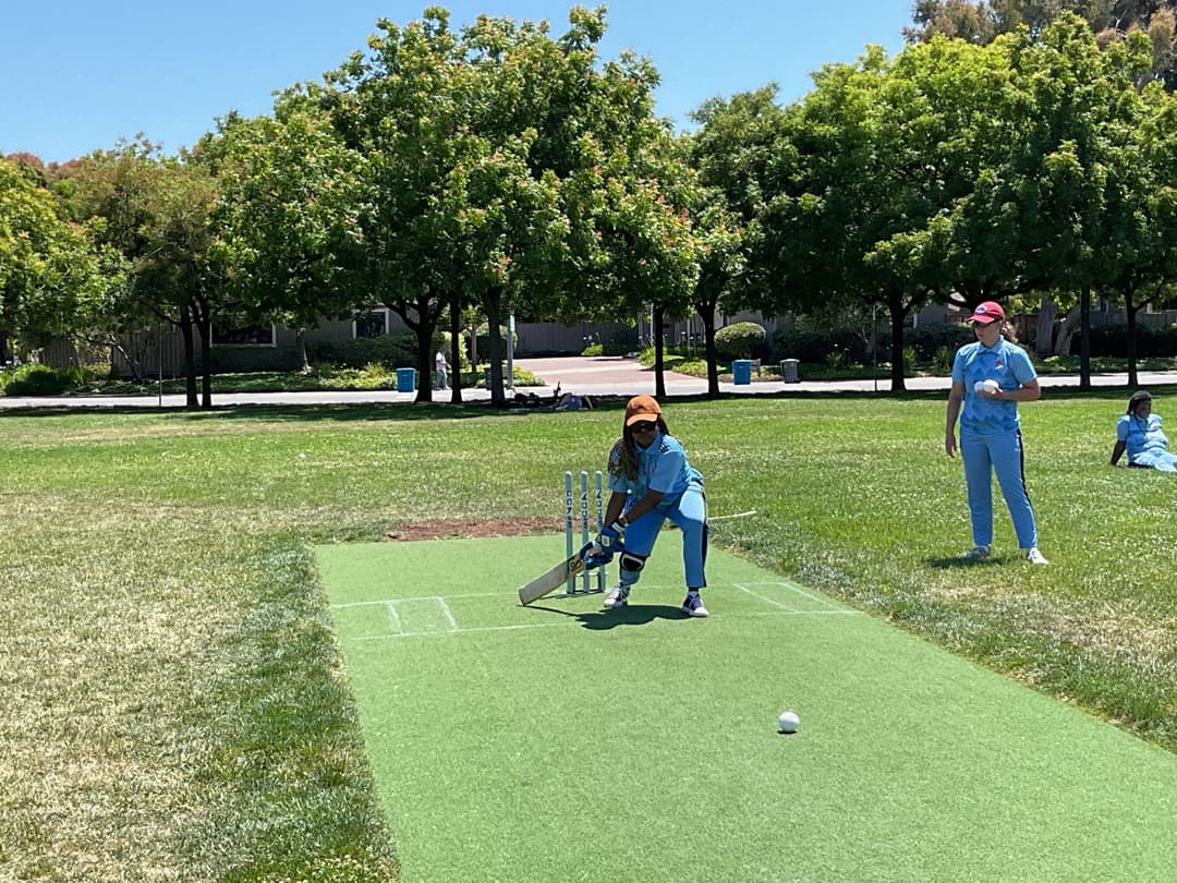 A bowler in the action to bowl, the ball has left her hand. The non striker, couple of fielders and the umpire are also visible in the shot. 