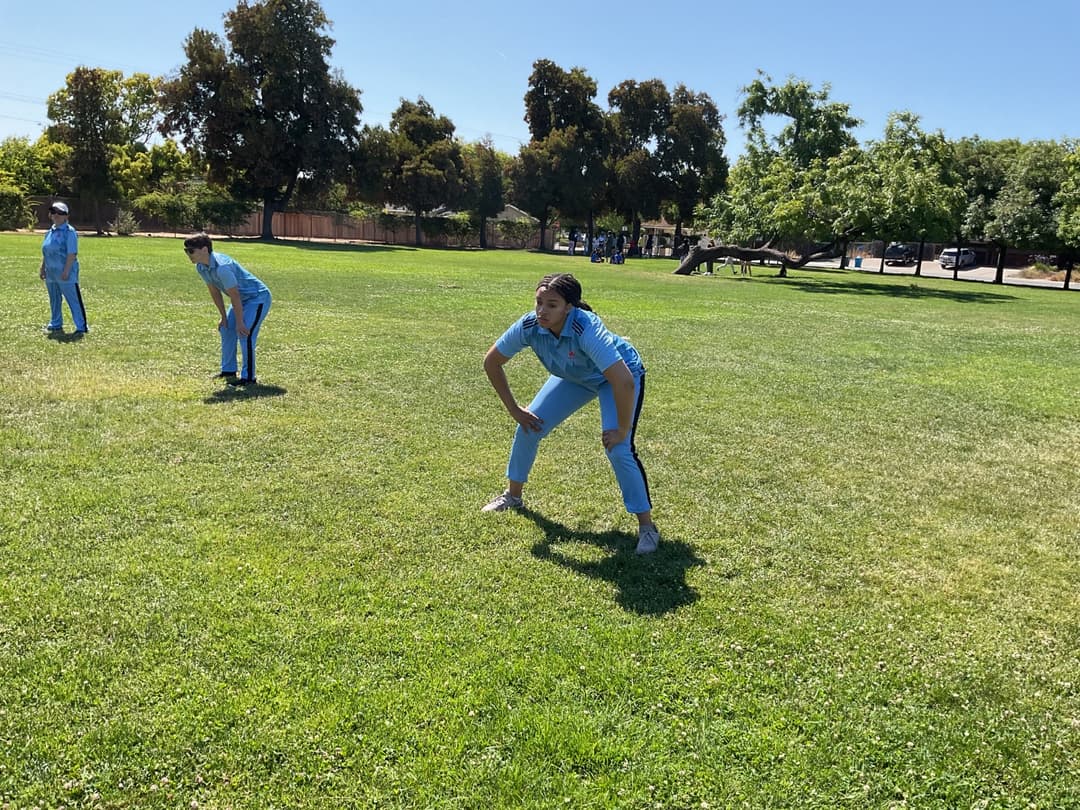 A bowler in the action to bowl, the ball has left her hand. The non striker, couple of fielders and the umpire are also visible in the shot. 