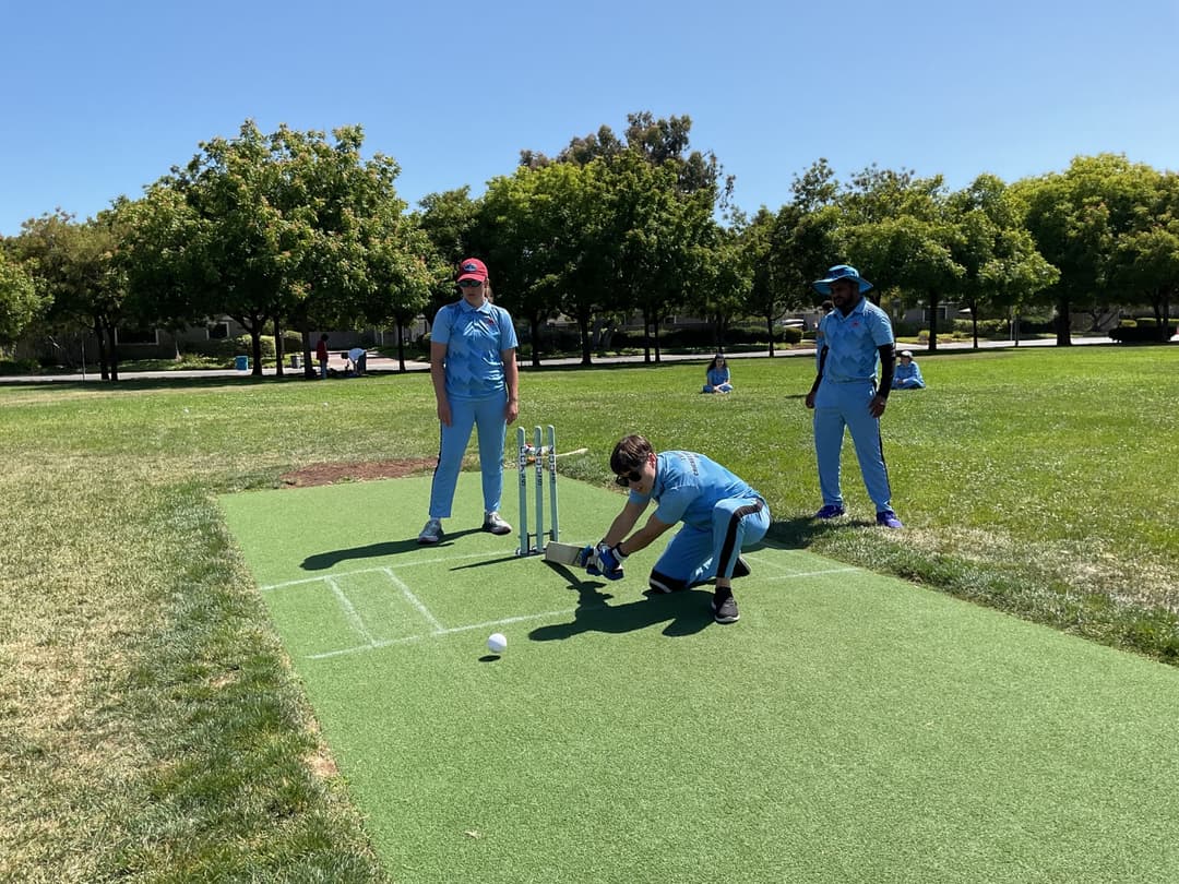 A bowler in the action to bowl, the ball has left her hand. The non striker, couple of fielders and the umpire are also visible in the shot. 
