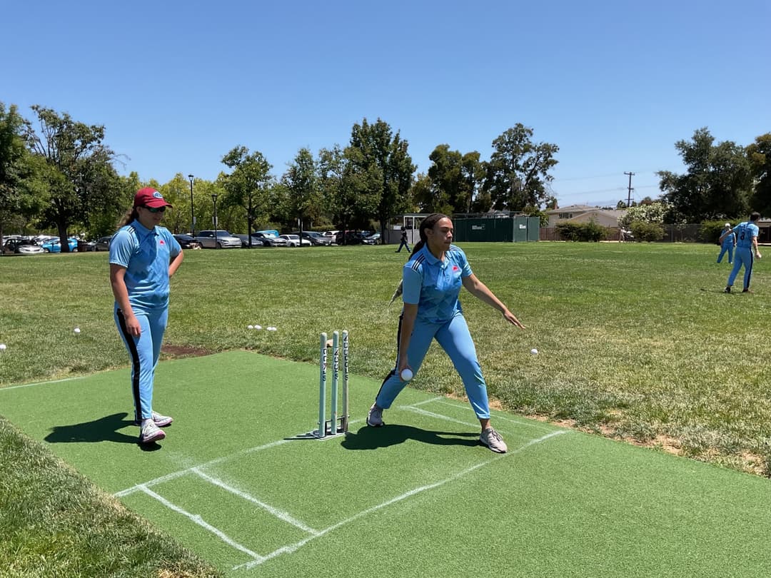 A bowler in the action to bowl, the ball has left her hand. The non striker, couple of fielders and the umpire are also visible in the shot. 