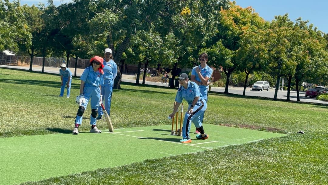 A bowler in the action to bowl, the ball has left her hand. The non striker, couple of fielders and the umpire are also visible in the shot. 
