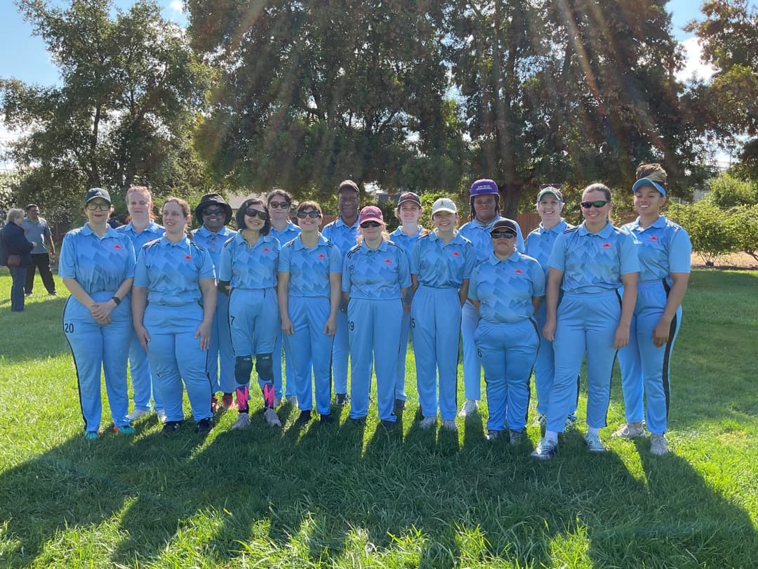 A bowler in the action to bowl, the ball has left her hand. The non striker, couple of fielders and the umpire are also visible in the shot. 