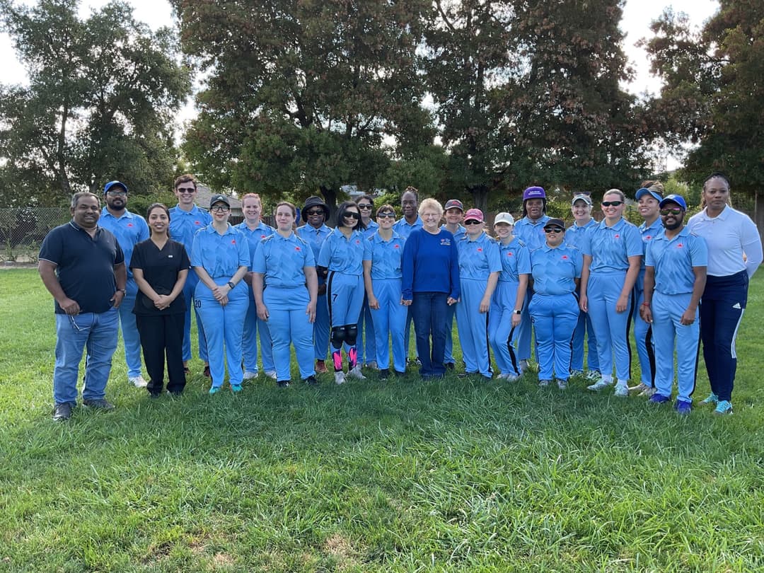 A bowler in the action to bowl, the ball has left her hand. The non striker, couple of fielders and the umpire are also visible in the shot. 