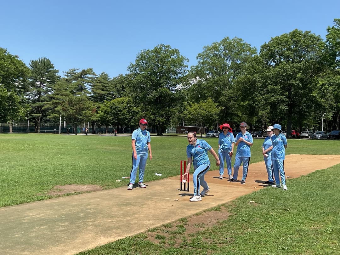 A bowler in the action to bowl, the ball has left her hand. The non striker, couple of fielders and the umpire are also visible in the shot. 