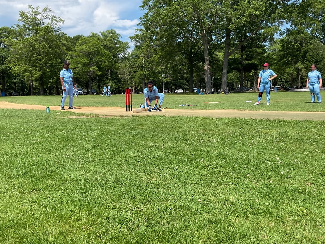 A bowler in the action to bowl, the ball has left her hand. The non striker, couple of fielders and the umpire are also visible in the shot. 